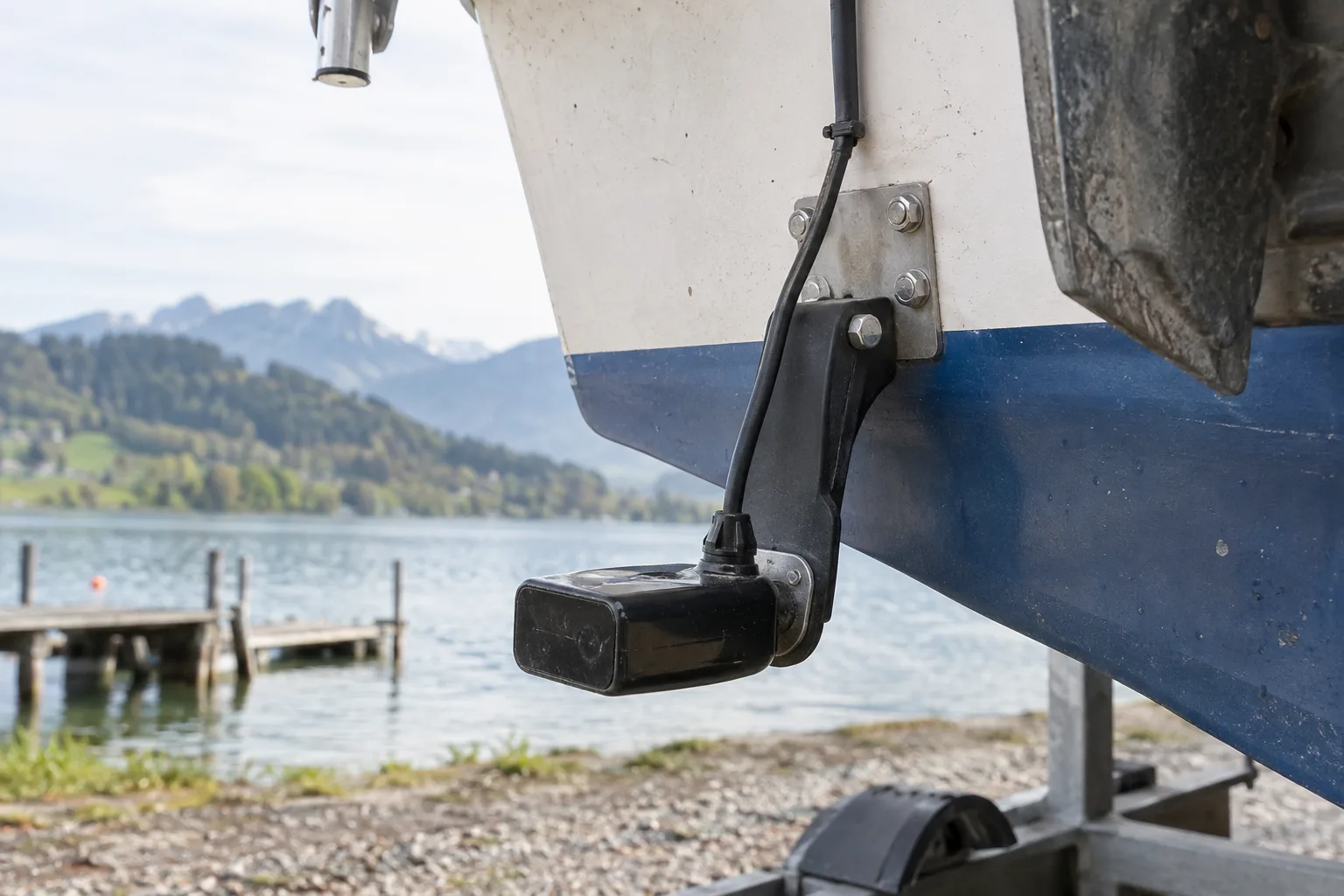 Echolot-Geber an der Heckspiegel-Halterung eines Bootes, verschraubte Montageplatte und nach oben geführtes Kabel; das Boot steht auf einem Trailer am Ufer.